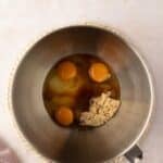 A metal mixing bowl containing three eggs, brown sugar, and vanilla extract sits on a counter with a mauve cloth nearby.