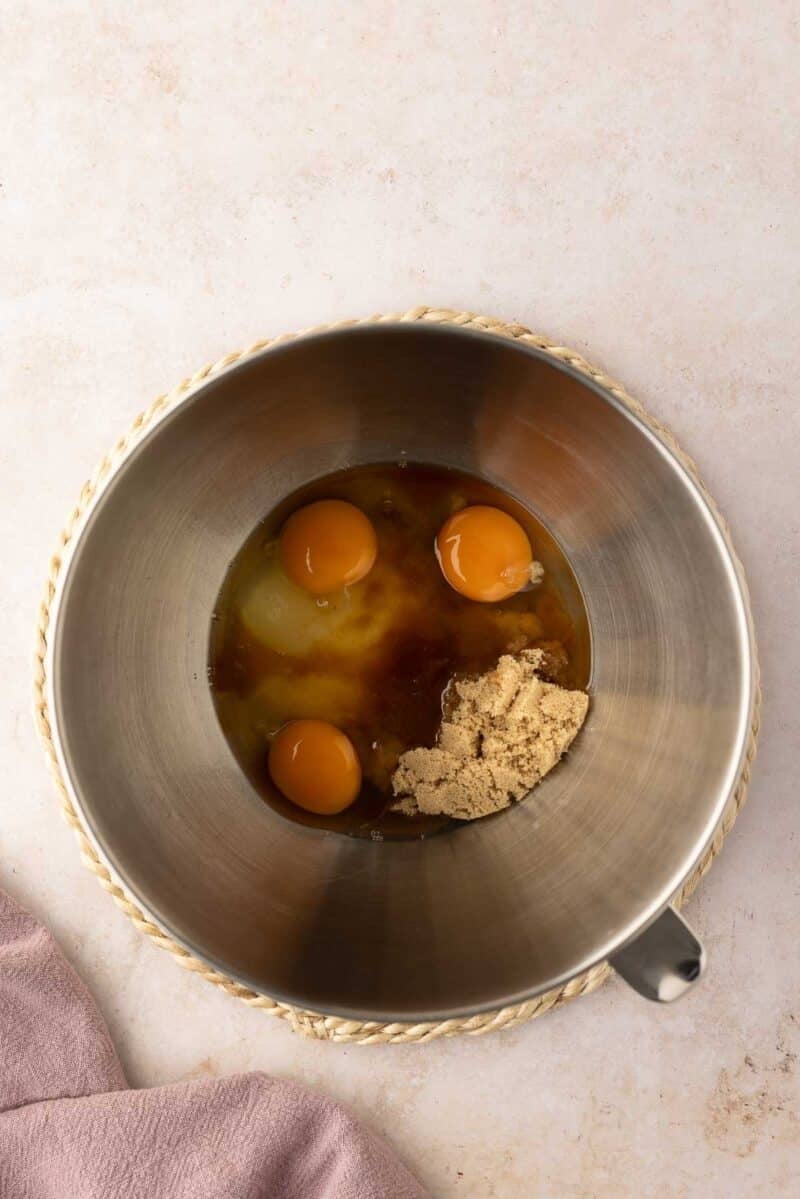 A metal mixing bowl containing three eggs, brown sugar, and vanilla extract sits on a counter with a mauve cloth nearby.