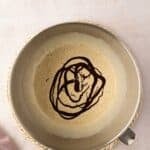 A metal mixing bowl with a light brown batter and chocolate sauce drizzled on top, placed on a woven mat next to a mauve cloth.