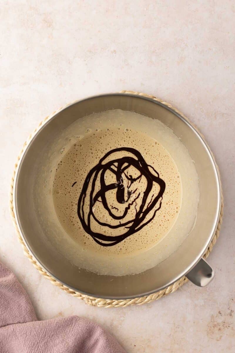 A metal mixing bowl with a light brown batter and chocolate sauce drizzled on top, placed on a woven mat next to a mauve cloth.