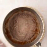 A metal mixing bowl with cocoa powder and flour mixture sits on a braided trivet, next to a light pink cloth on a beige surface.
