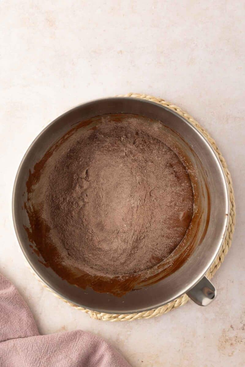 A metal mixing bowl with cocoa powder and flour mixture sits on a braided trivet, next to a light pink cloth on a beige surface.