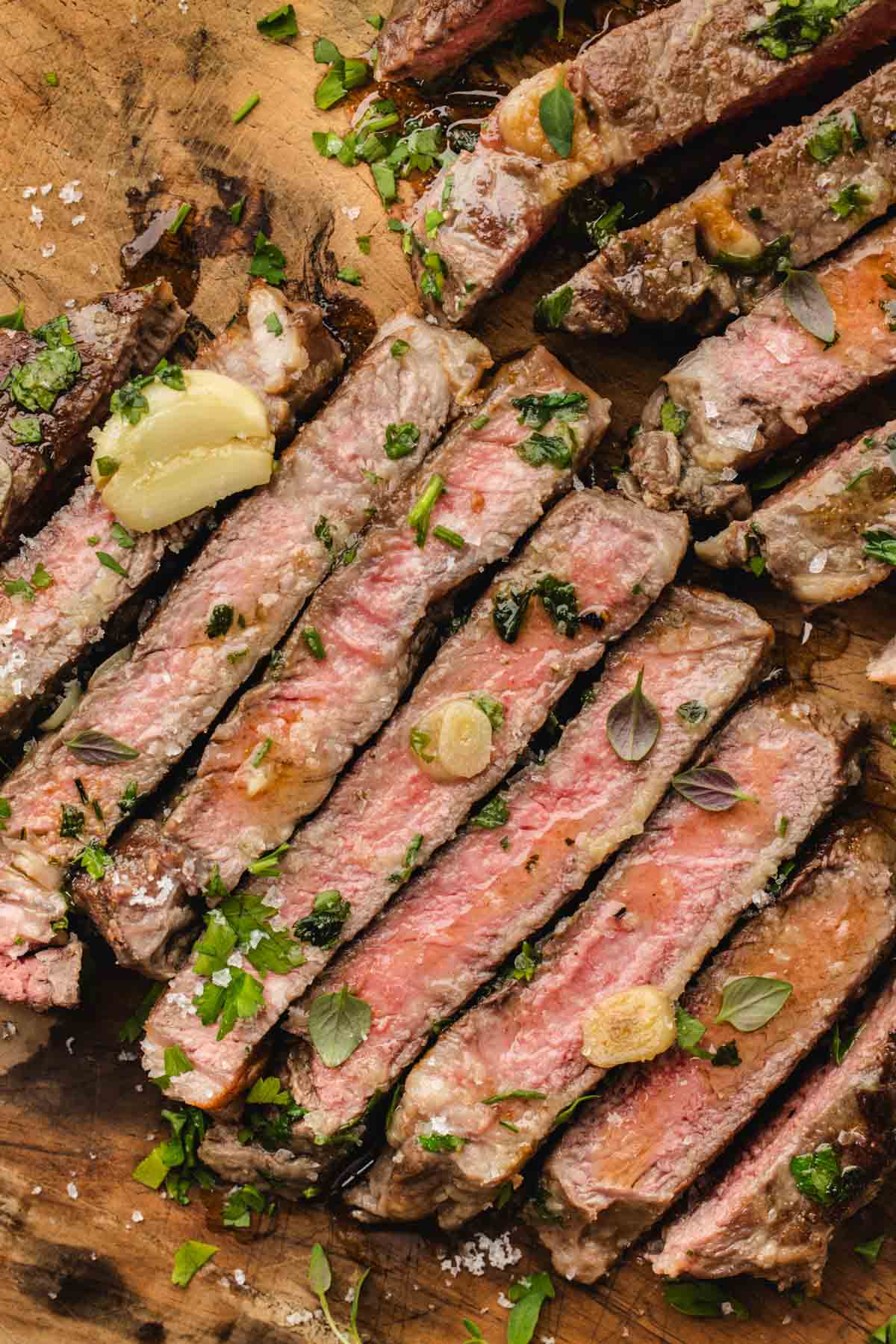 Sliced medium-rare ribeye steak topped with herbs and garlic on a wooden cutting board.