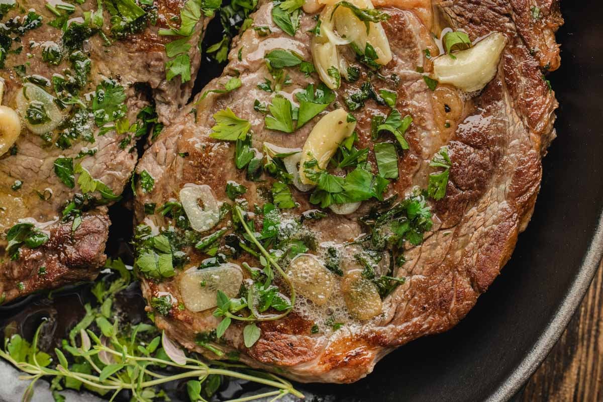 Close-up of cooked ribeye steak in a pan, topped with chopped parsley, sliced garlic, and melted butter.