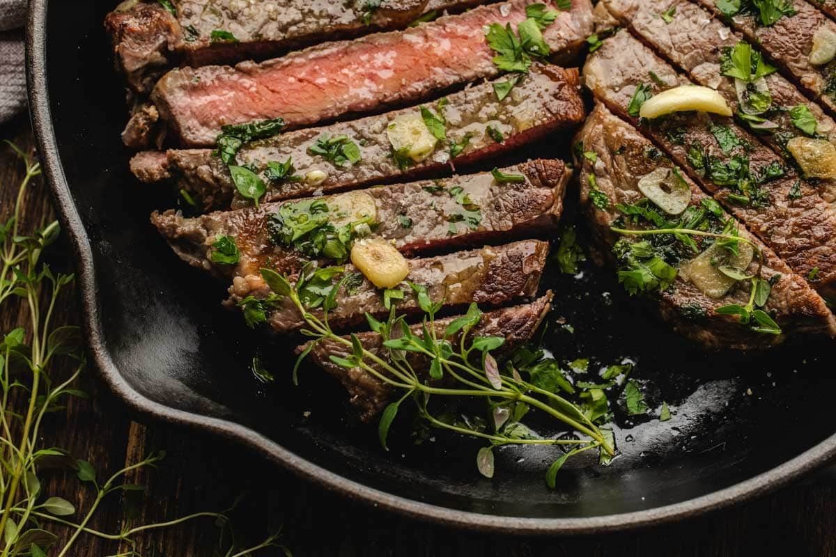 Sliced steak cooked medium rare in a cast iron skillet, topped with fresh herbs and garlic slices.