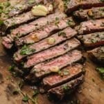 Sliced medium-rare ribeye steak on a wooden board, topped with herbs, sliced garlic, and a pat of butter.