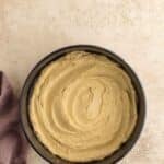 A round cake pan filled with smooth, swirled cake batter sits on a textured surface next to a folded mauve cloth.