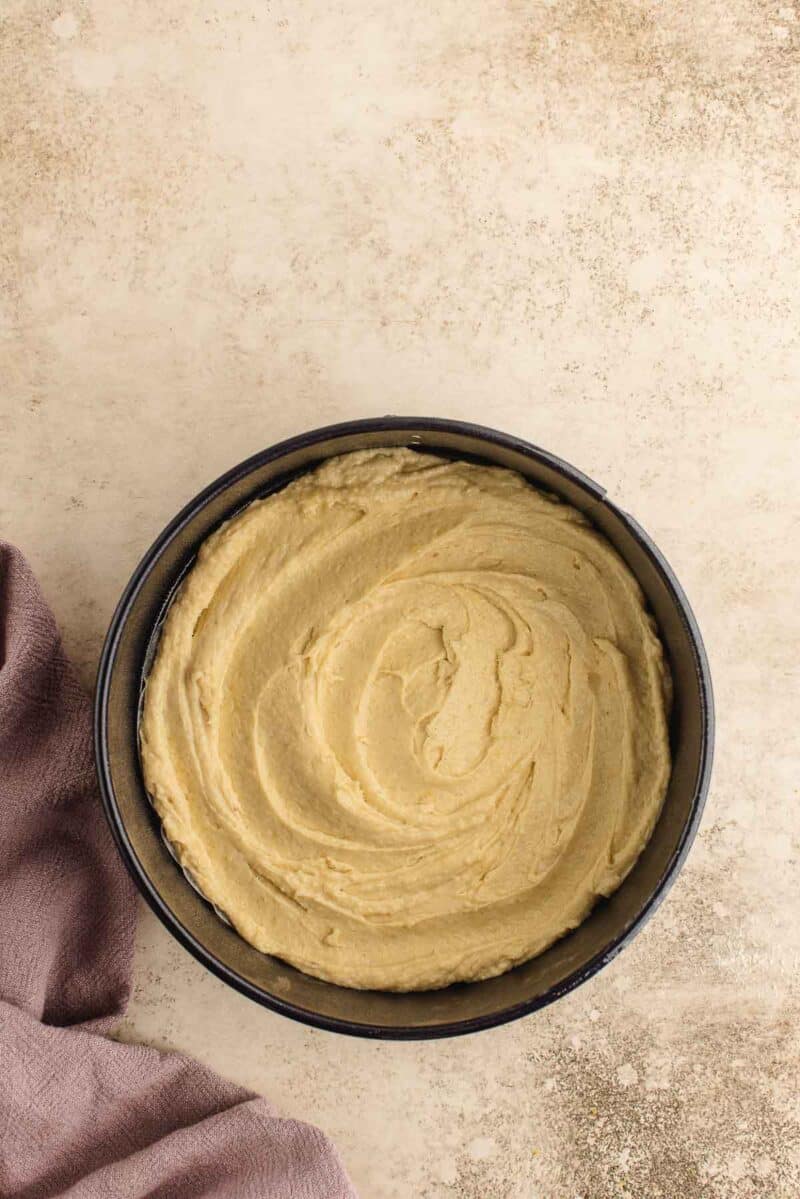 A round cake pan filled with smooth, swirled cake batter sits on a textured surface next to a folded mauve cloth.