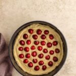 A round cake pan filled with unbaked batter, topped with evenly spaced cherries, sits on a light surface next to a folded mauve cloth.