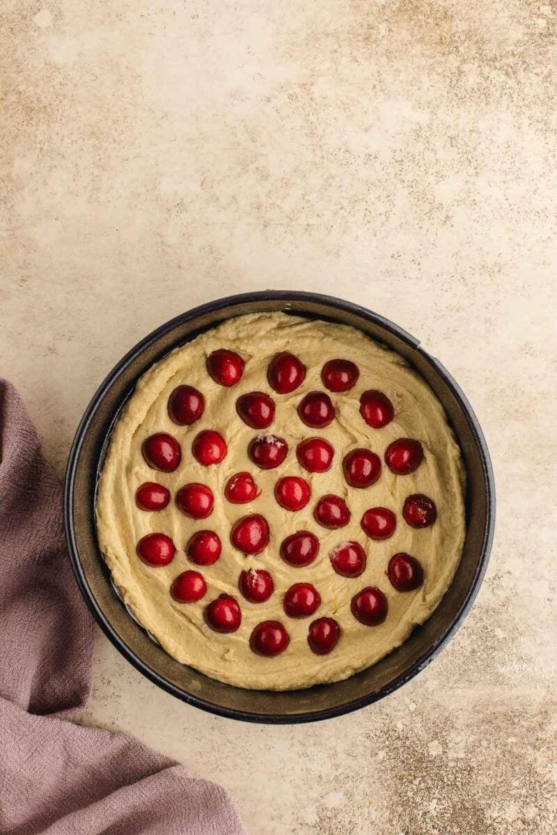 A round cake pan filled with unbaked batter, topped with evenly spaced cherries, sits on a light surface next to a folded mauve cloth.