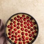 A round baking pan filled with batter topped with evenly spaced red cherries sits on a light countertop beside a folded purple cloth.