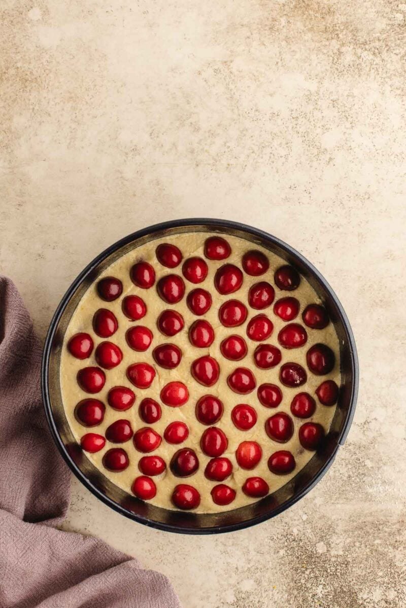 A round baking pan filled with batter topped with evenly spaced red cherries sits on a light countertop beside a folded purple cloth.