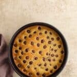 A round cake with evenly spaced chocolate chips on top sits in a black baking pan on a light textured surface next to a folded purple cloth.