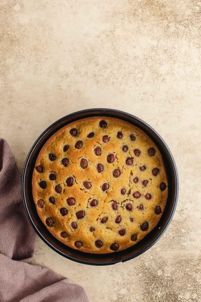 A round cake with evenly spaced chocolate chips on top sits in a black baking pan on a light textured surface next to a folded purple cloth.