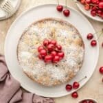 Round cake dusted with powdered sugar, topped with fresh cherries, surrounded by more cherries on a light surface with a mauve cloth and plates of forks.