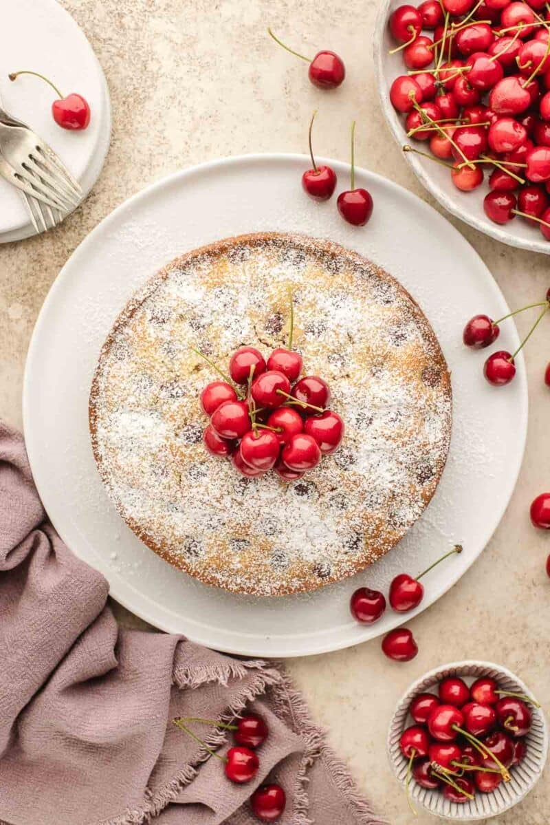 Round cake dusted with powdered sugar, topped with fresh cherries, surrounded by more cherries on a light surface with a mauve cloth and plates of forks.