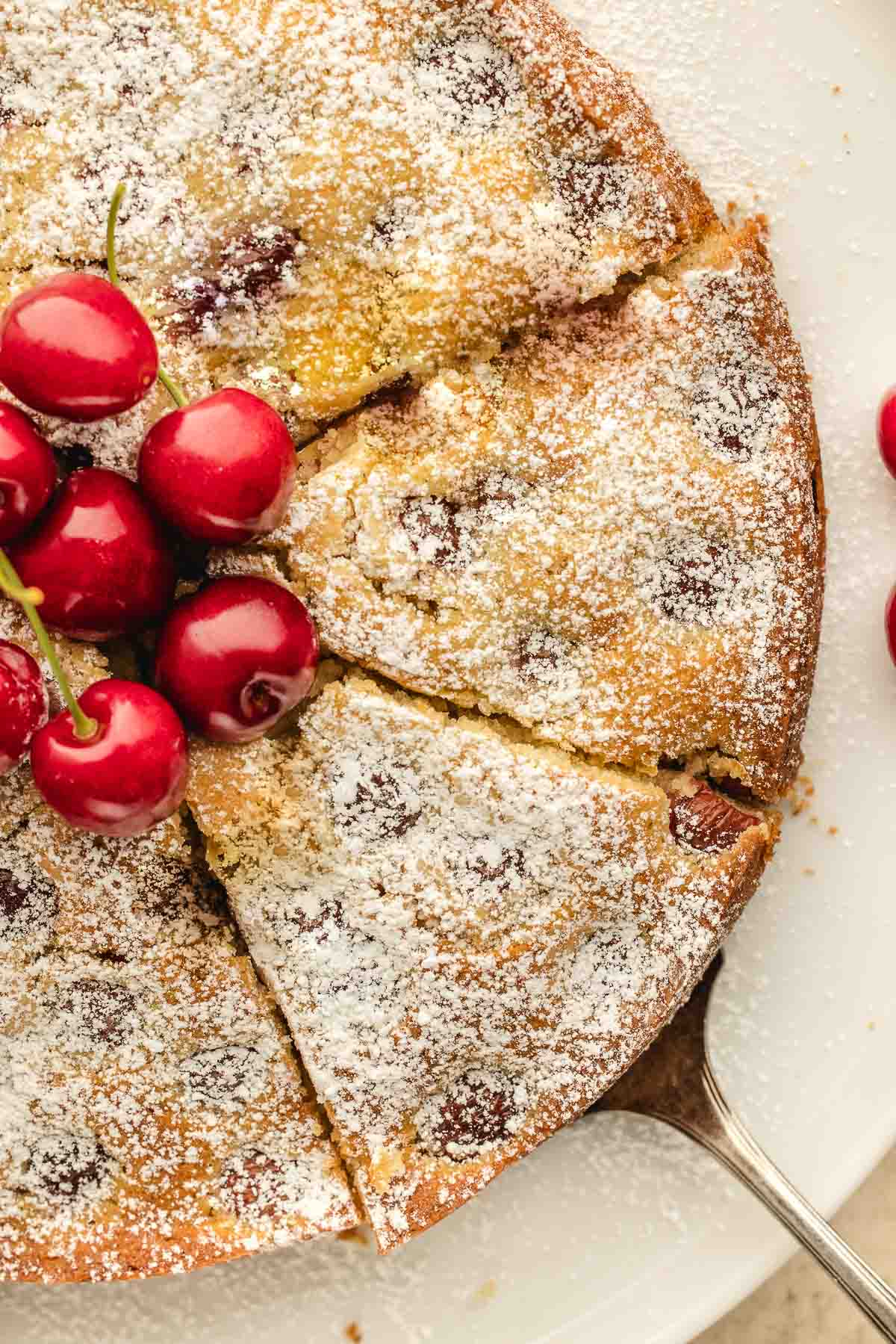 A round cherry cake topped with powdered sugar and whole cherries, sliced and served on a white plate with a cake server.