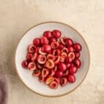 A white bowl filled with halved pitted cherries sits on a light textured surface.