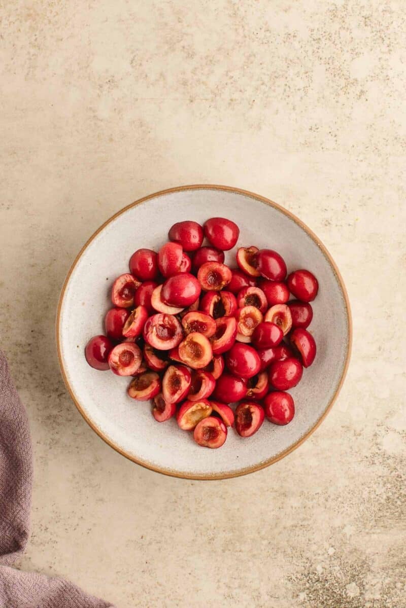 A white bowl filled with halved pitted cherries sits on a light textured surface.