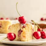 A slice of cherry cake garnished with fresh cherries on a white plate, with more cake and cherries in the background.