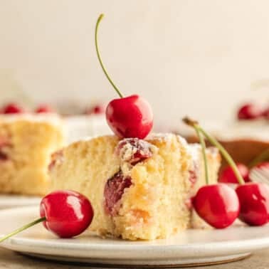 A slice of cherry cake garnished with fresh cherries on a white plate, with more cake and cherries in the background.