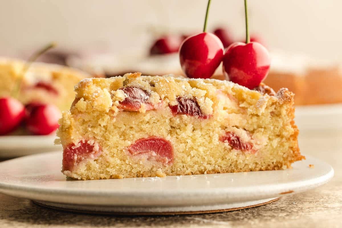 A slice of cherry almond cake with visible cherry pieces sits on a plate, with two whole cherries on top and more cherries blurred in the background.