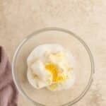 A glass bowl with sugar, butter, and lemon zest sits on a light textured surface next to a folded mauve cloth.