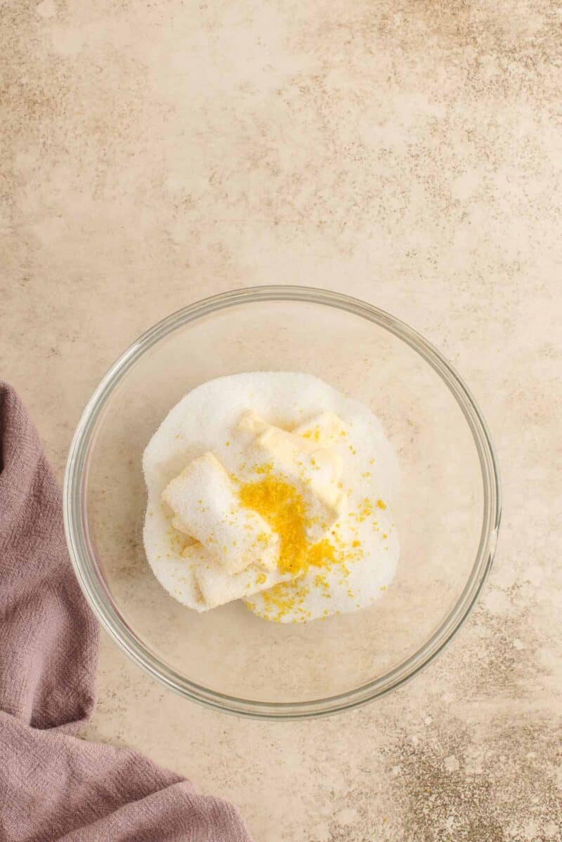 A glass bowl with sugar, butter, and lemon zest sits on a light textured surface next to a folded mauve cloth.