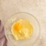 A glass bowl with creamed butter and sugar, plus one raw egg on top, sits on a light countertop next to a folded lavender cloth.