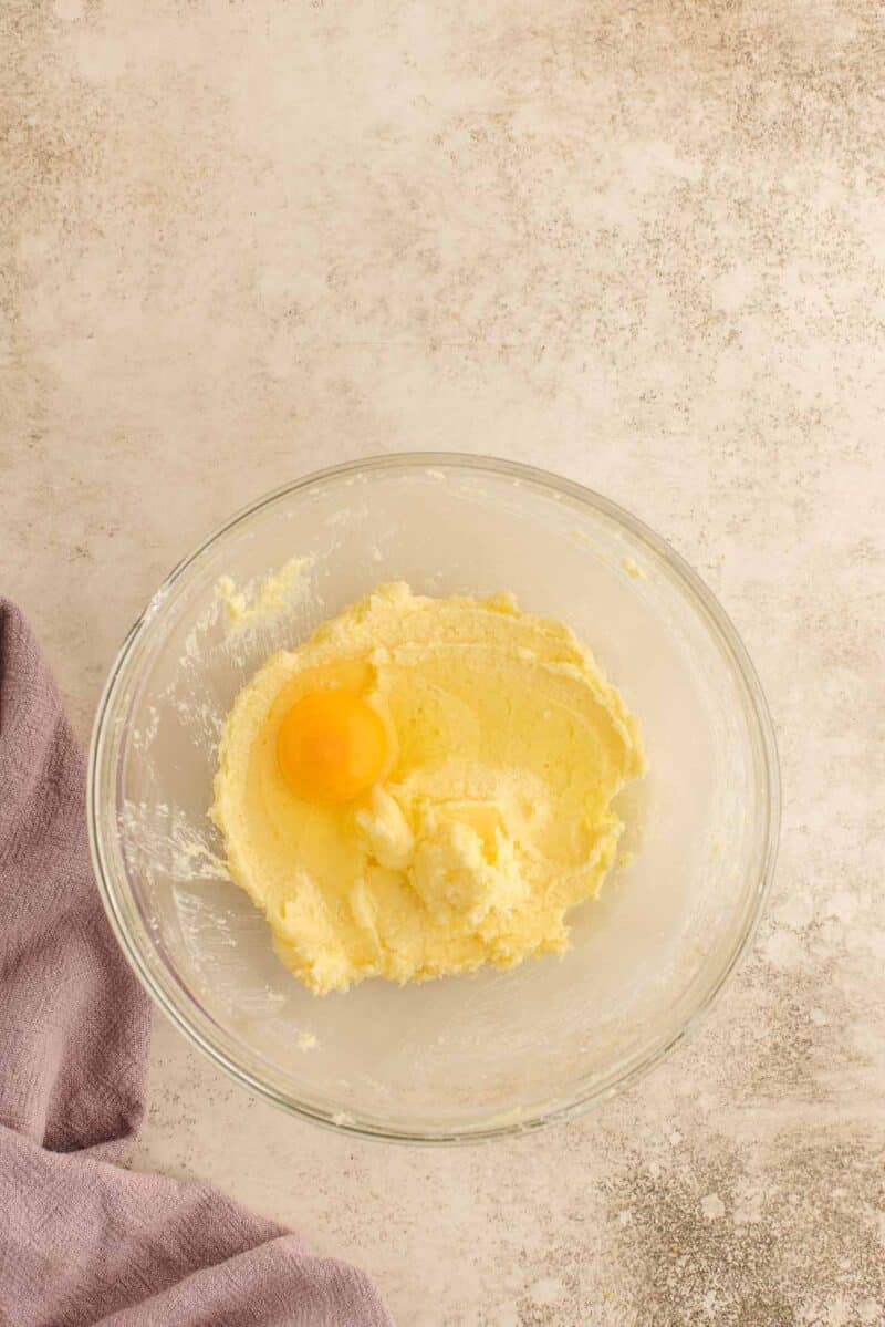 A glass bowl with creamed butter and sugar, plus one raw egg on top, sits on a light countertop next to a folded lavender cloth.