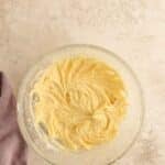 A glass mixing bowl filled with creamed cake batter sits on a light textured surface next to a folded lavender dish towel.