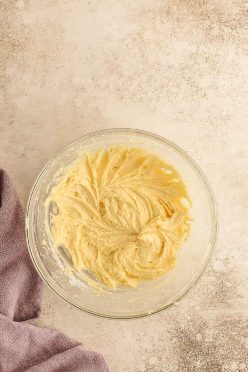 A glass mixing bowl filled with creamed cake batter sits on a light textured surface next to a folded lavender dish towel.