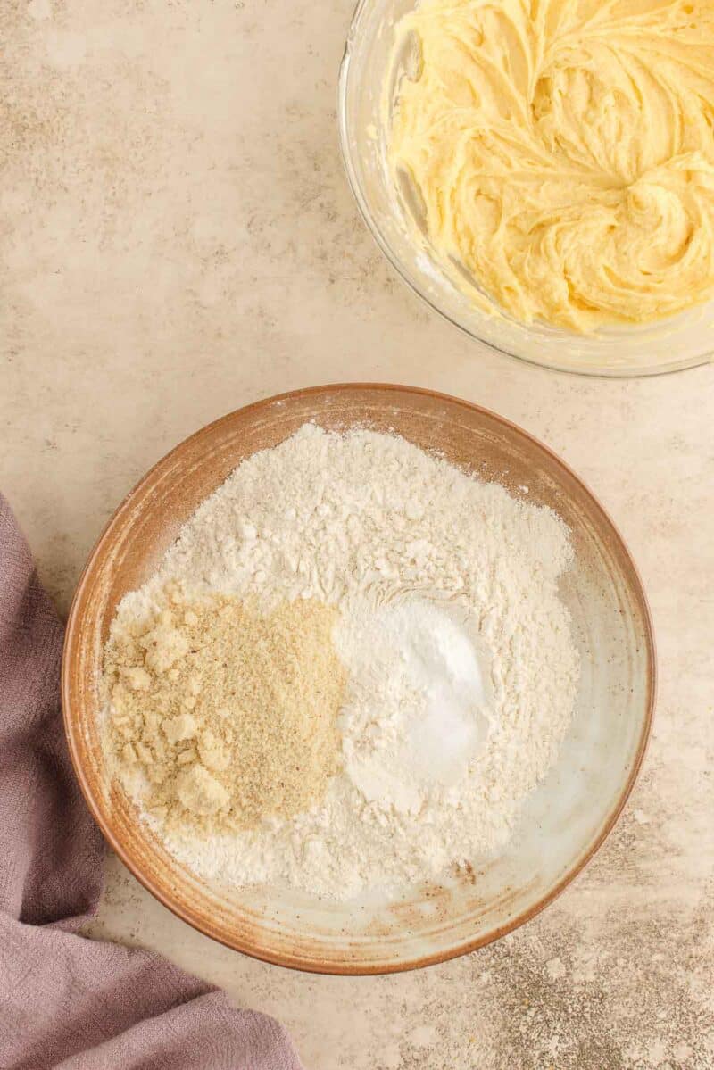 A bowl with flour, almond flour, baking powder, and salt next to a bowl of creamed butter and sugar on a textured surface.