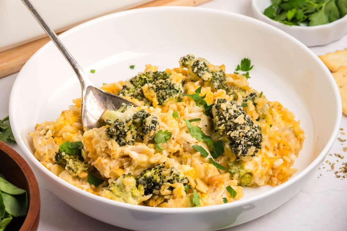A white bowl filled with a chicken broccoli rice casserole topped with parsley, with a spoon resting inside. Crackers and fresh herbs are visible in the background.