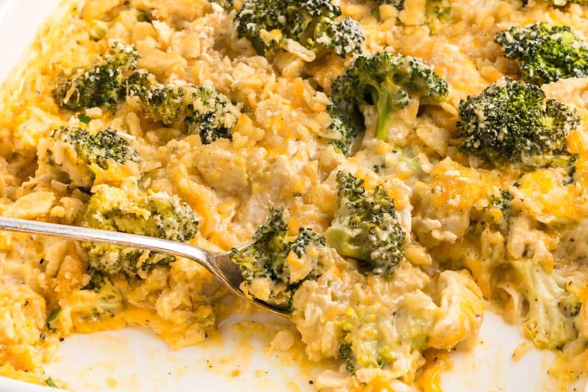 A close-up of a cheesy broccoli and chicken casserole in a baking dish, with a serving spoon scooping out a portion.