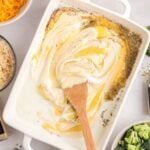 A wooden spoon stirs creamy sauce with seasoning in a white baking dish, surrounded by bowls of shredded cheese, chicken, broccoli, crackers, and rice.