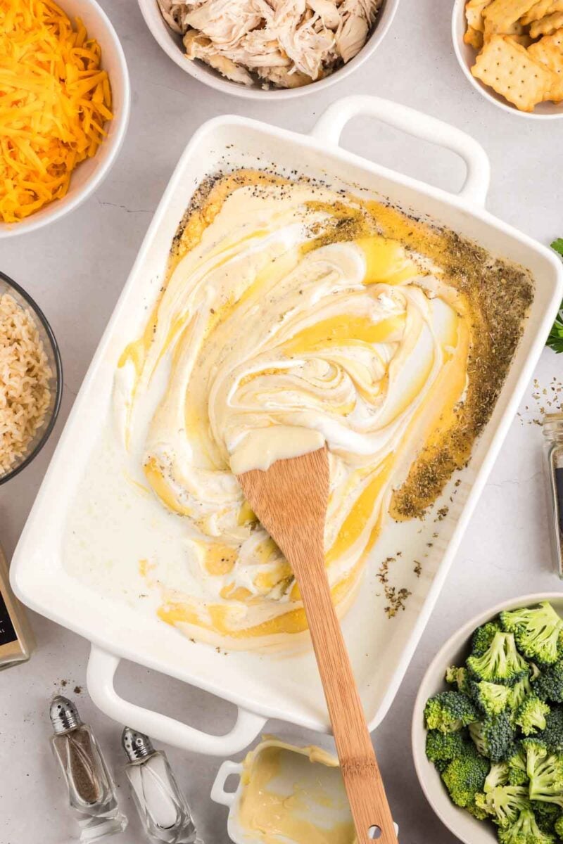 A wooden spoon stirs creamy sauce with seasoning in a white baking dish, surrounded by bowls of shredded cheese, chicken, broccoli, crackers, and rice.