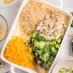 A casserole dish with shredded chicken, cooked rice, broccoli florets, and shredded cheddar cheese being mixed with a wooden spoon.