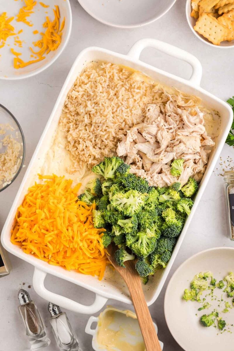 A casserole dish with shredded chicken, cooked rice, broccoli florets, and shredded cheddar cheese being mixed with a wooden spoon.