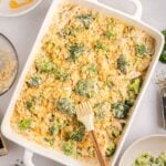 A casserole dish filled with broccoli and chicken topped with crushed crackers, surrounded by bowls of cheese, crackers, and kitchen utensils on a light surface.