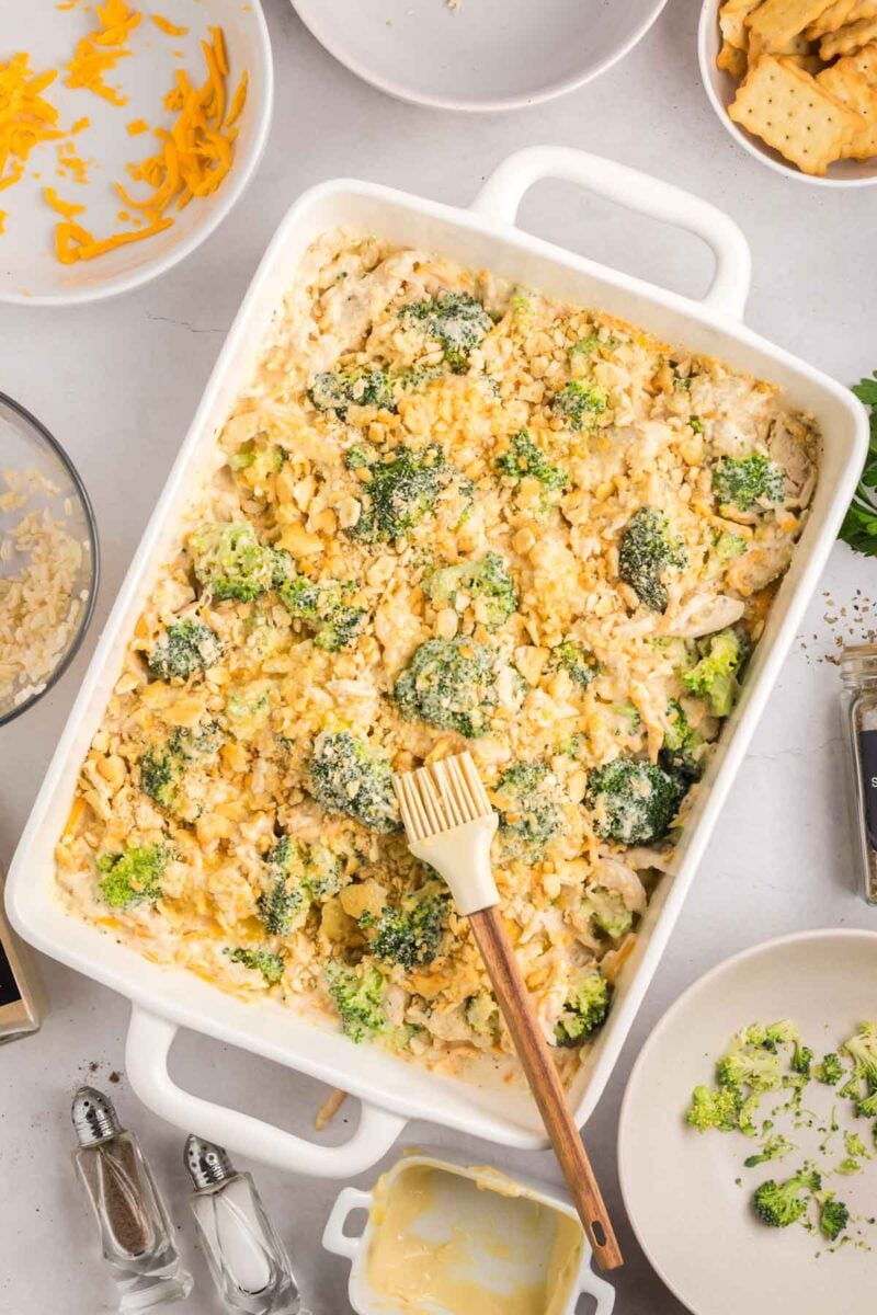 A casserole dish filled with broccoli and chicken topped with crushed crackers, surrounded by bowls of cheese, crackers, and kitchen utensils on a light surface.