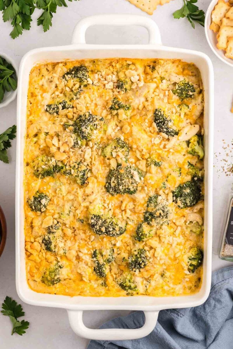 A baked casserole in a white dish, featuring chicken broccoli rice casserole, and a golden cracker topping, surrounded by parsley and crackers on a light surface.