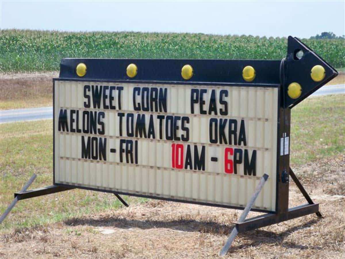 Roadside sign advertises sweet corn, peas, melons, tomatoes, and okra for sale, available Monday to Friday from 10 AM to 6 PM.