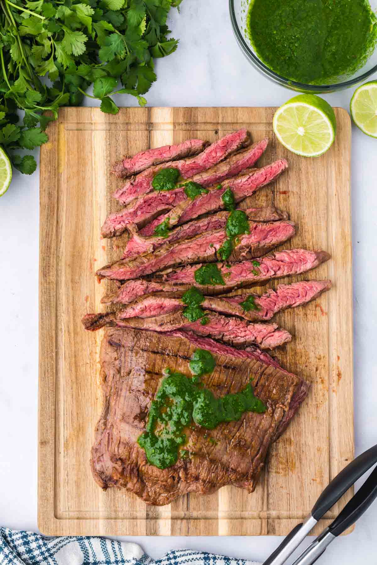 Sliced grilled flank steak with green chimichurri sauce on a wooden cutting board, garnished with lime halves and cilantro, with metal tongs and a bowl of sauce nearby.