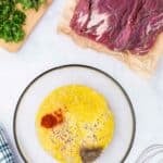 Raw flank steak on parchment next to a cutting board with fresh herbs and a bowl of beaten eggs with spices, with a whisk and kitchen towel nearby on a white surface.