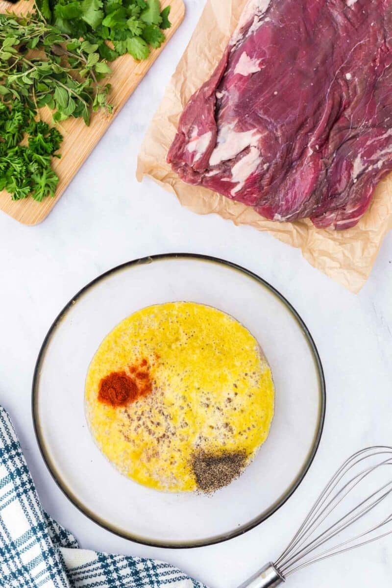 Raw flank steak on parchment next to a cutting board with fresh herbs and a bowl of beaten eggs with spices, with a whisk and kitchen towel nearby on a white surface.