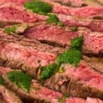 Sliced medium-rare flank steak on a wooden board, topped with dollops of green chimichurri sauce.