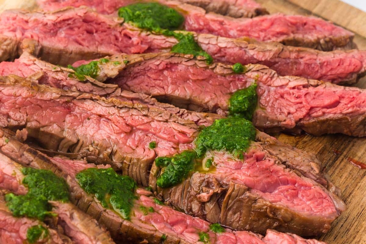 Sliced medium-rare flank steak on a wooden board, topped with dollops of green chimichurri sauce.