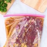 Raw steak marinating in a sealed plastic bag with herbs and cutting boards nearby on a white surface.