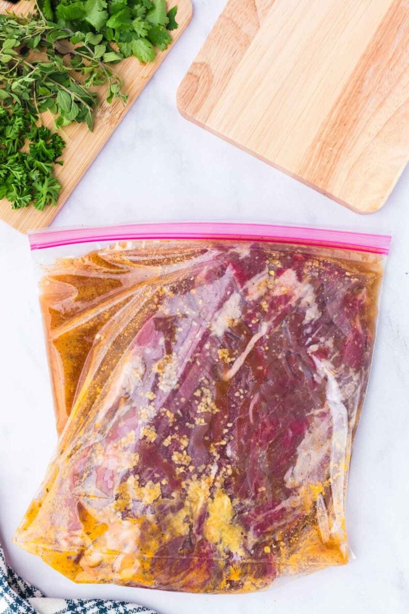 Raw steak marinating in a sealed plastic bag with herbs and cutting boards nearby on a white surface.